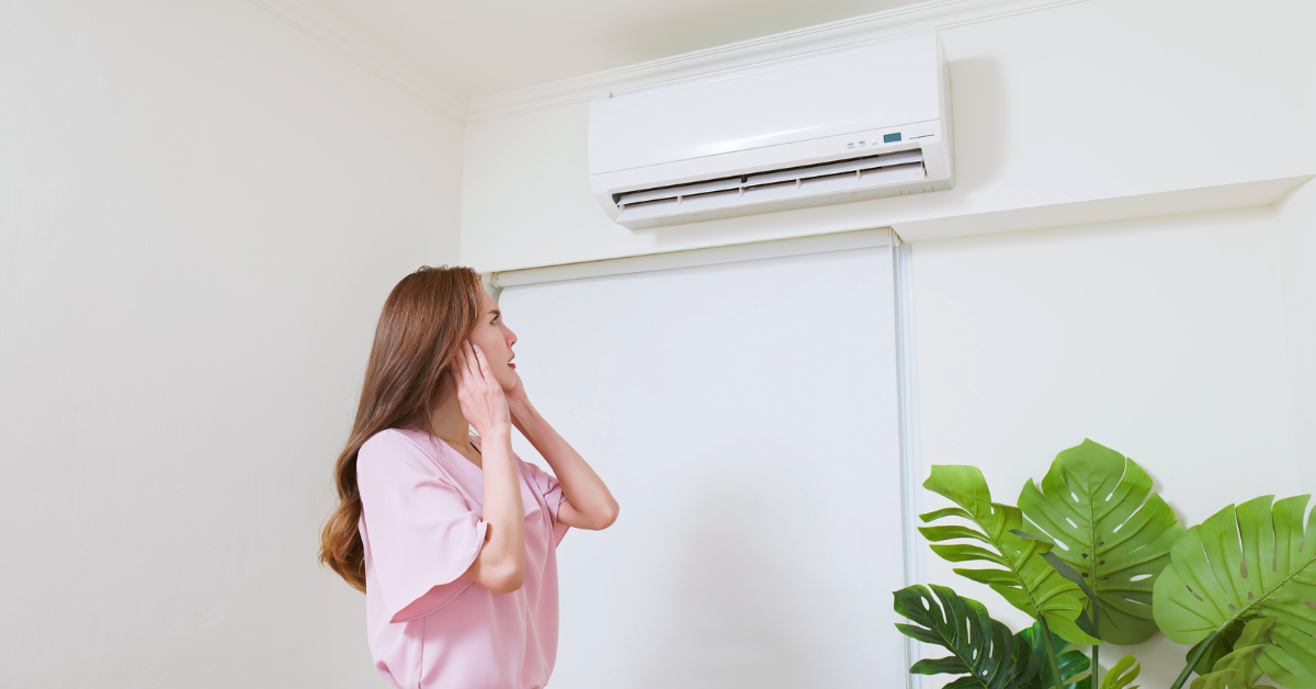 A woman protects her ears and looks up at a noisy air conditioner